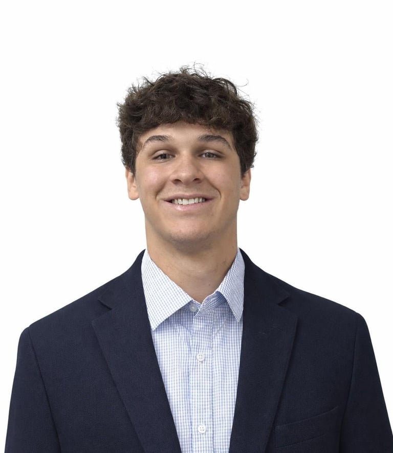 Young man with dark curly hair smiling at camera, wearing navy blazer and light blue checkered shirt